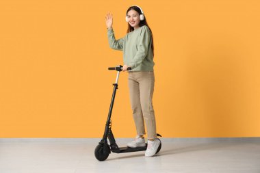 Sarı duvarın yanında modern elektrikli scooterlı, kulaklıklı güzel bir Asyalı kadın.