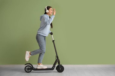 Kulaklıklı güzel bir Asyalı kadın yeşil duvarın yanında modern elektrikli scooter kullanıyor.