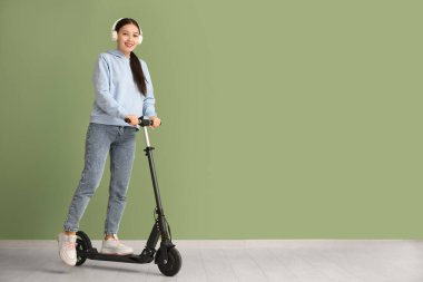 Yeşil duvarın yanında modern elektrikli scooterlı kulaklıklı güzel bir Asyalı kadın.