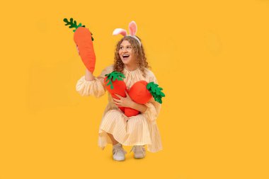 Tavşan kulaklı, sarı arka planda havuç şeklinde oyuncakları olan genç ve güzel bir kadın. Paskalya kutlaması