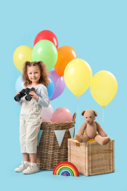 Elinde dürbün, balonlu sepetler ve mavi arka planda oyuncaklar tutan sevimli küçük bir kız.