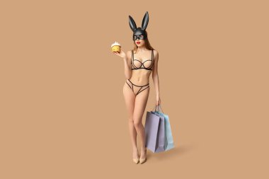 Alışveriş torbaları ve bej arka planda Paskalya pastası olan tavşan maskeli seksi genç kadın.