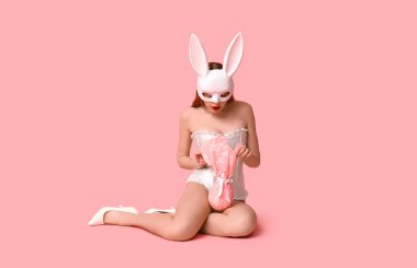 Paskalya hediyeli, pembe arka planda oturan tavşan maskeli seksi genç kadın.