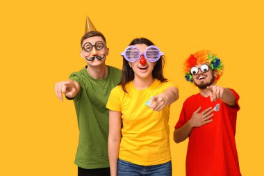 Sarı arka plandaki izleyiciyi işaret eden kağıt balıklarıyla komik kılık değiştirmiş genç arkadaşlar. 1 Nisan kutlamaları