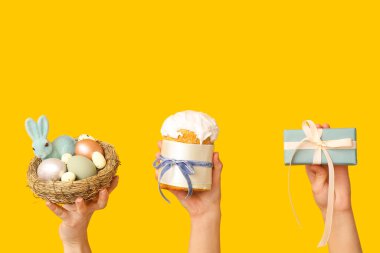 Yumurtalı yuva, paskalya pastası ve sarı arka planda hediye kutusu olan kadın elleri.