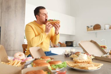 Mutfaktaki sağlıksız yiyeceklerle dolu masada sosisli sandviç yiyen genç bir adam. Aşırı yeme kavramı