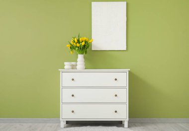 Çekmece dolabında sarı lale vazosu ve oturma odasındaki duvarda resim.