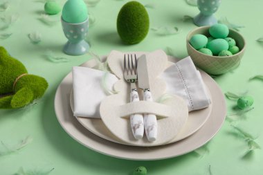 Peçeteli tabaklar, çatal bıçak takımı, paskalya yumurtalı kase ve yeşil arka planda tüyler.