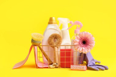 İçinde temizlik malzemeleri, aletler ve sarı arka planda bahar çiçeği olan sepet.