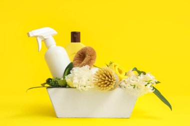 İçinde temizlik malzemeleri, aletler ve sarı arka planda bahar çiçekleri olan sepet.