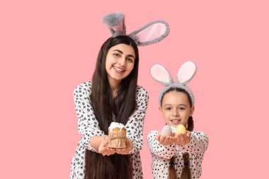 Mutlu gülümseyen genç bir kadın, kızıyla tavşan kulaklı, elinde paskalya pastası ve pembe arka planda yumurtalar.