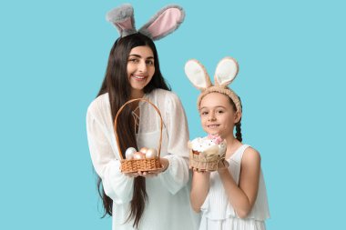 Mutlu gülümseyen genç bir kadın, kızıyla tavşan kulaklı, elinde paskalya pastası ve mavi arka planda yumurtaları olan bir sepet tutuyor.