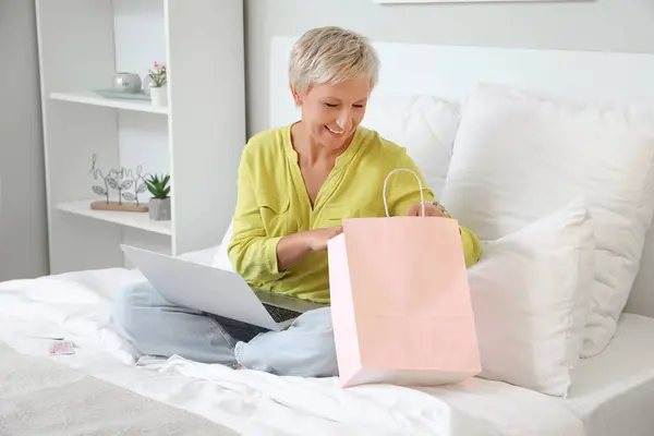 Mature woman with laptop and shopping bag in bedroom