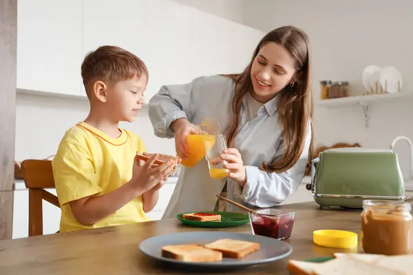 Mother pouring juice for her little son with toasts in kitchen