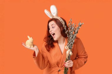Surprised young woman in bunny ears with pussy willow branches and toy rabbit on orange background. Easter celebration