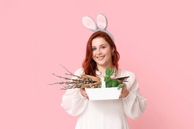 Beautiful young woman in bunny ears with pussy willow branches and Easter basket on pink background