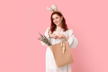 Beautiful young woman in bunny ears holding paper bag with pussy willow branches on pink background. Easter celebration