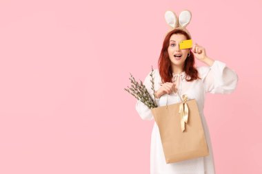 Beautiful young woman in bunny ears holding paper bag with pussy willow branches and credit card on pink background. Easter celebration