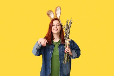Beautiful young woman in bunny ears with pussy willow branches on yellow background. Easter celebration