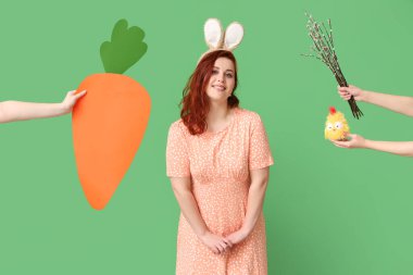 Beautiful young woman in bunny ears and hands with pussy willow branches, paper carrot and toy chick on green background. Easter celebration
