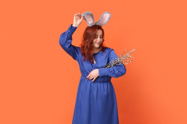 Beautiful young woman in bunny ears with pussy willow branches on orange background. Easter celebration
