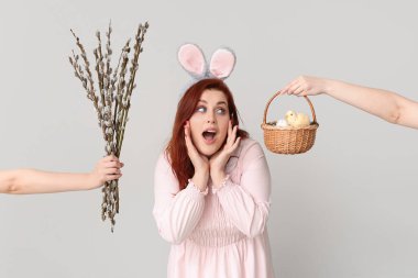 Shocked young woman in bunny ears and hands with pussy willow branches and Easter basket on grey background