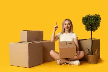 Karton kutuları ve çiçekleri olan mutlu genç bir kadın sarı arka plandaki bir şeyi işaret ediyor. Yeni ev konseptine geçiyoruz.