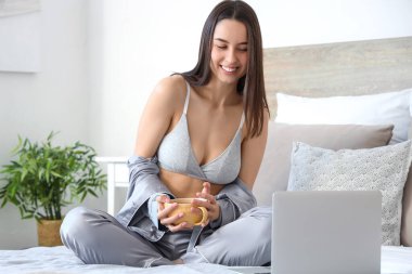 Yatak odasında çay içen dizüstü bilgisayarı olan seksi genç bir kadın.