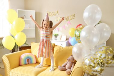 Parti şapkasıyla kanepede oturan tatlı küçük kız evde doğum günü için balonlar ve süslemelerle.