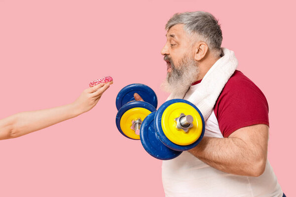 Избыточный взрослый мужчина с гантелями и пончиками на розовом фоне. Концепция потери веса