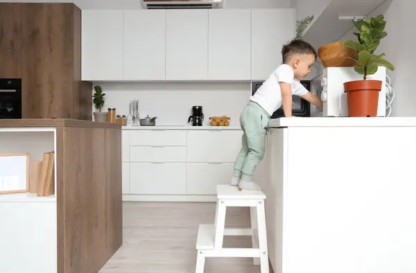 Little boy on step stool opening microwave oven in kitchen. Child at risk