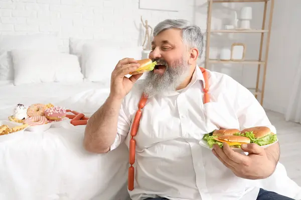 Overweight mature man eating burgers in bedroom. Overeating concept