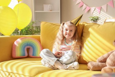 Parti şapkasıyla kanepede oturan sevimli küçük kız balonlarla, oyuncaklarla ve evde doğum günü süsleriyle.