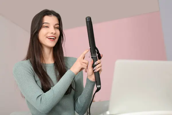 Beautiful young happy female blogger with laptop and flattening iron recording video at home