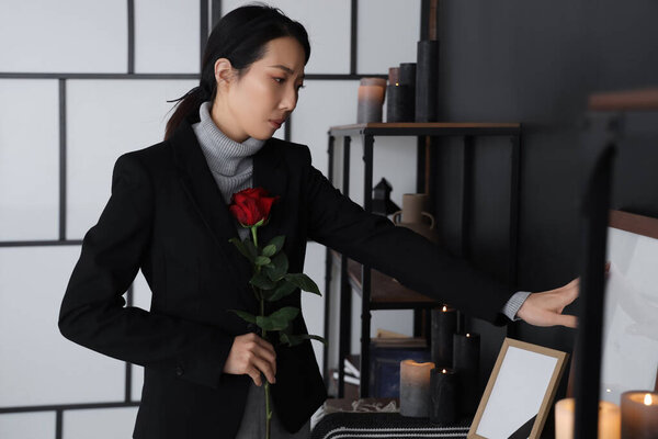 Mourning young Asian woman with rose near photo frame at funeral