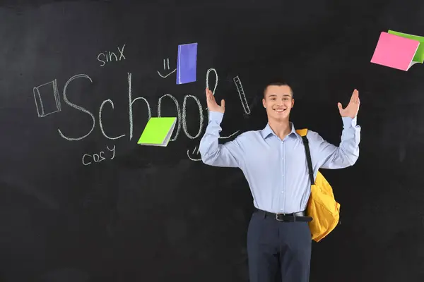 Male student throwing copybooks away near word SCHOOL written on blackboard. End of school concept