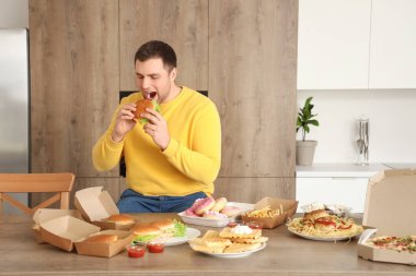 Mutfaktaki sağlıksız yiyeceklerle dolu masada hamburger yiyen genç bir adam. Aşırı yeme kavramı