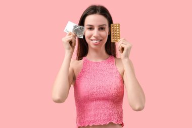 Pembe arka planda doğum kontrol hapları ve prezervatifleri olan güzel bir genç kadın.
