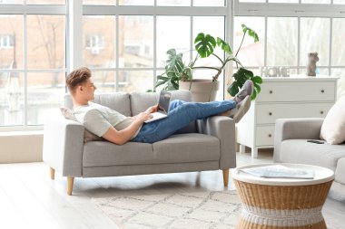 Evdeki kanepede dizüstü bilgisayar kullanan yakışıklı genç adam.