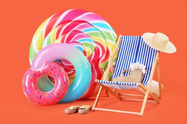 Şeker şeklinde yüzen yatak, şişme yüzük ve turuncu arka planda hasır şapkalı güneş şezlongu.