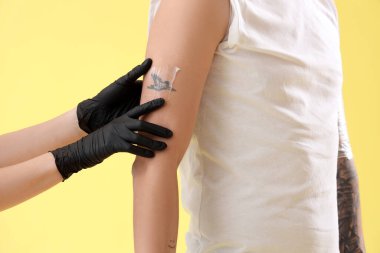 Dövme ustası adamın koluna krem sürüyor. Sarı arka plan, yakın plan.