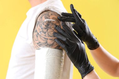 Dövme ustası adamın omzuna krem sürüyor. Sarı arka plan, yakın plan.