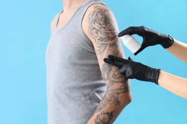 Dövme ustası adamın koluna krem sürüyor, mavi arka planda, yakın plan.
