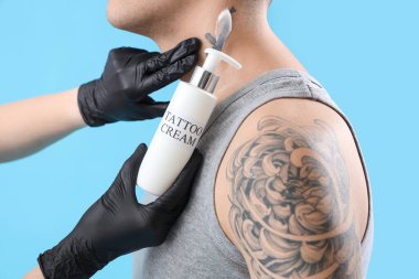 Dövme ustası, adamın boynuna krem sürüyor, mavi arka plan, yakın plan.