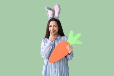 Paskalya tavşanı kulaklı, yeşil arka planda kağıt havuç olan güzel genç bir kadın.