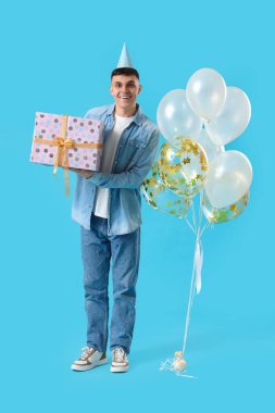 Mavi arka planda bir sürü balonu ve hediye kutusu olan genç bir adam.