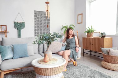 Oturma odasındaki kanepede oturan rüzgar çanı çalan genç kadın.