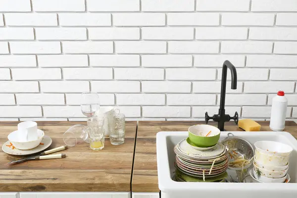 Wooden counter with ceramic sink and dirty dishes near white brick wall