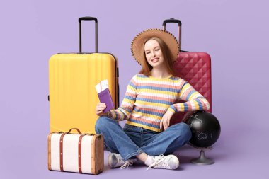 Pasaport ve küreyle saman şapkalı gülümseyen genç bir kadın leylak arka planında bavulunun yanında oturuyor. Seyahat kavramı