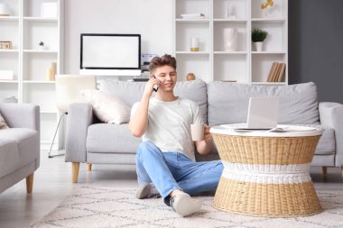 Bir fincan kahve ve dizüstü bilgisayarlı genç adam evde cep telefonuyla konuşuyor.